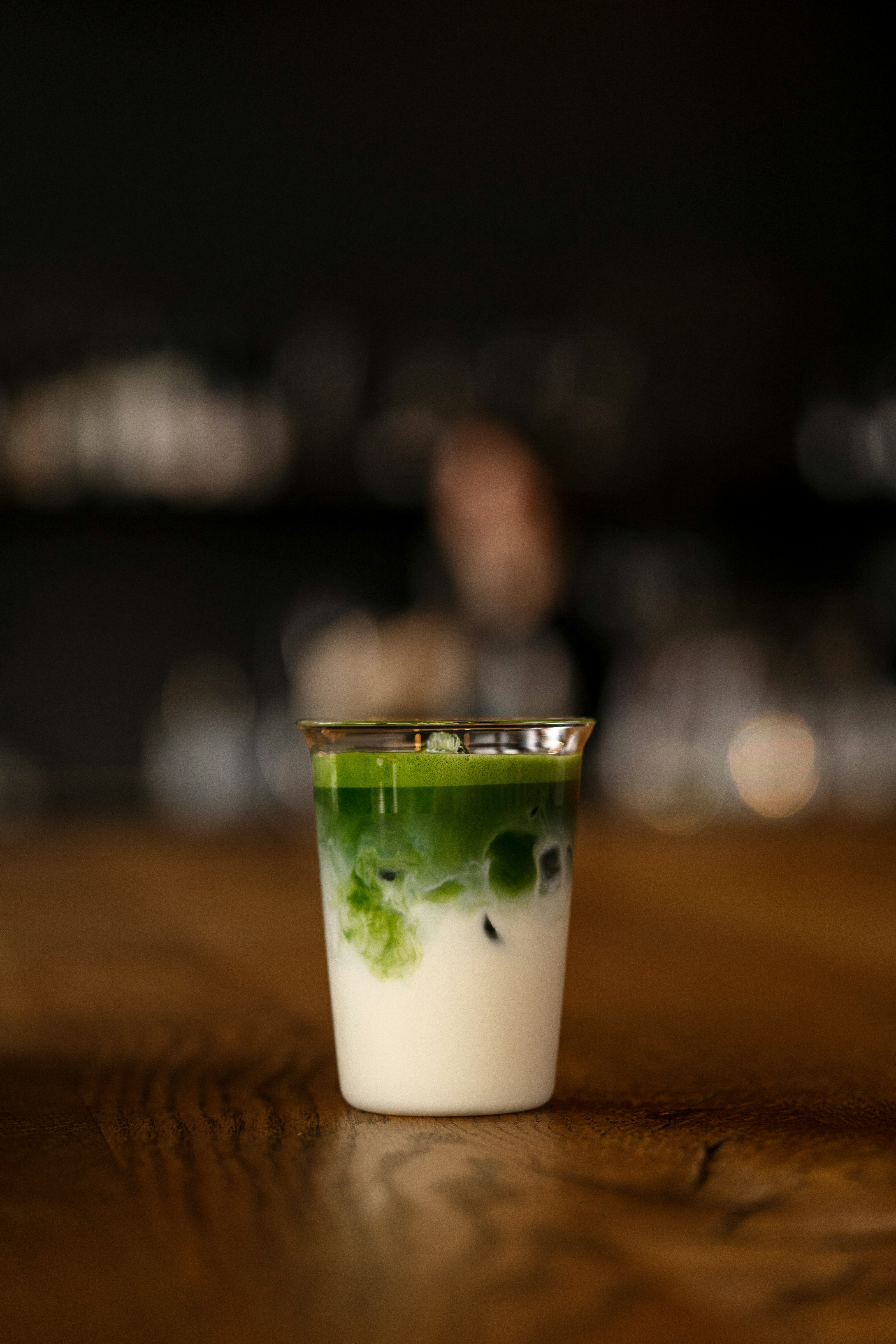 A matcha latte is being poured over milk in a glass on a wooden table, creating a layered green and white drink.